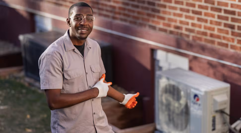 An HVAC worker standing in front of a heat pump giving a thumbs up and smiling