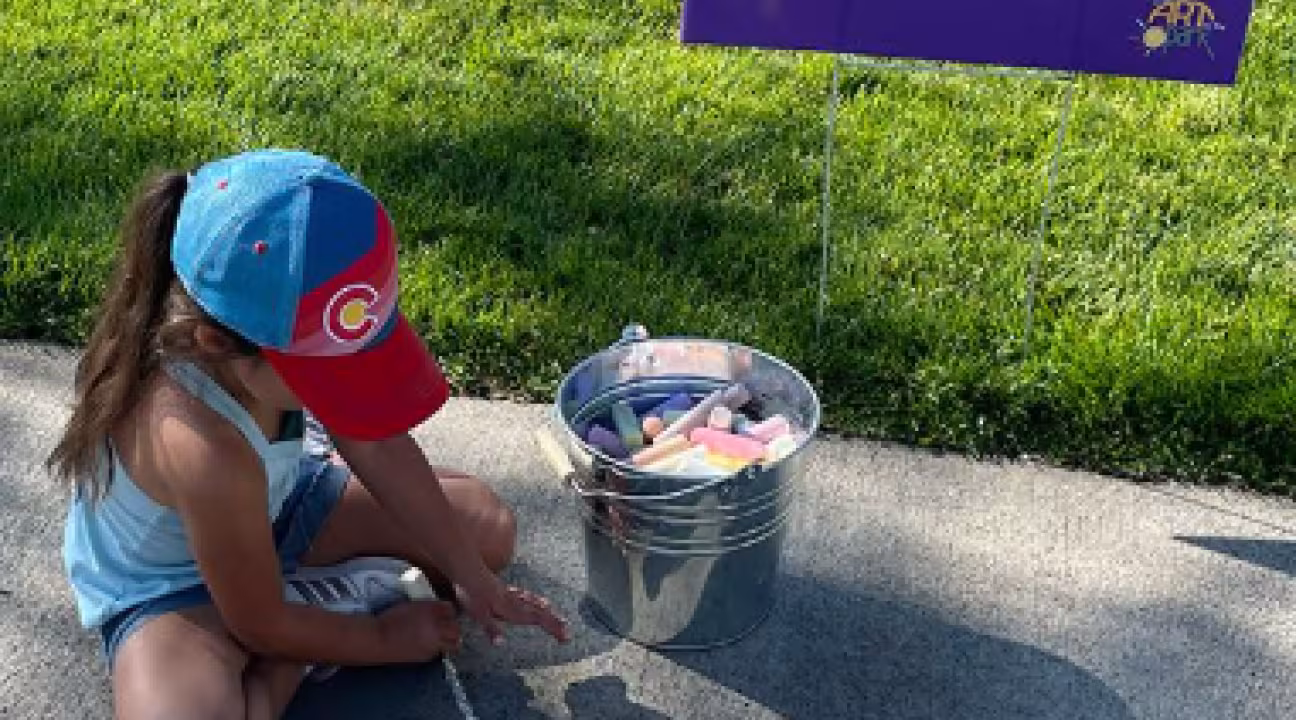 Small child drawing on the sidewalk in front of a sign that reads "Leave Your Mark Here!"